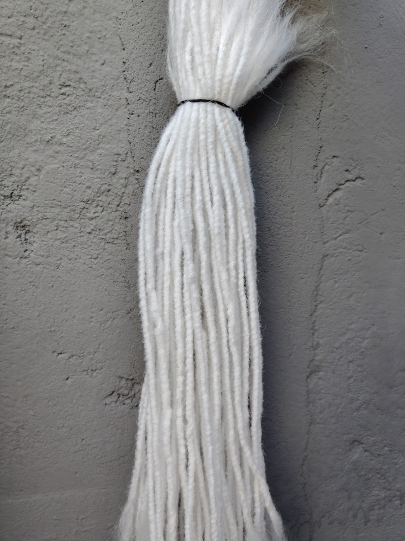 A close-up of a bundle of white yarn or dreadlocks tied with a black band against a textured gray wall.