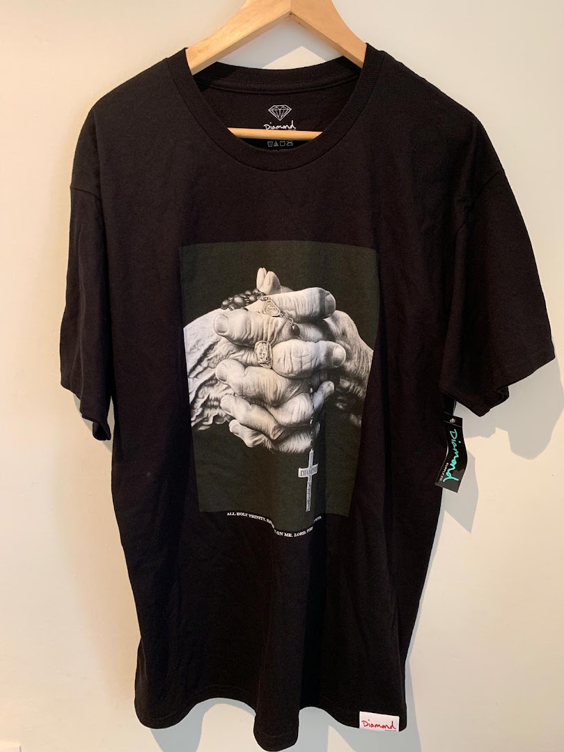 A black t-shirt with a graphic of praying hands and a rosary hangs on a wooden hanger.