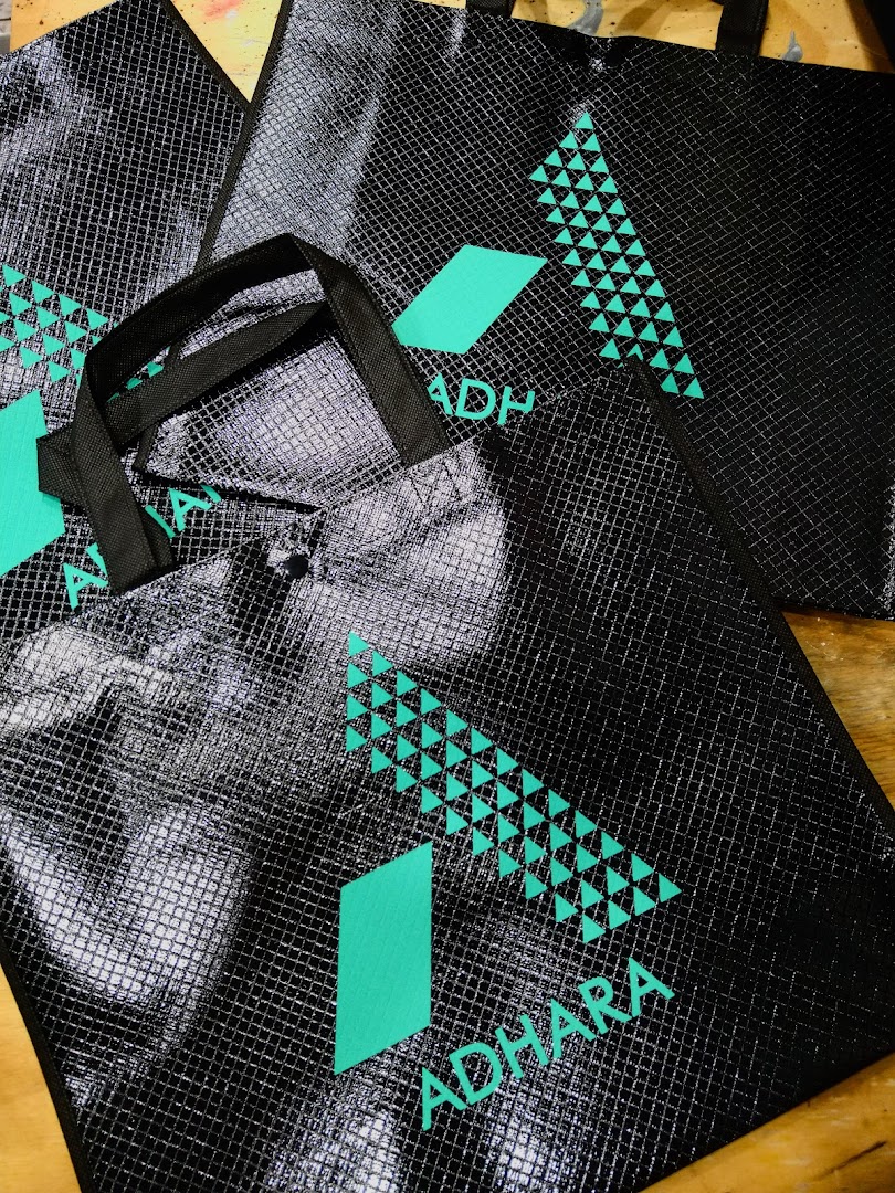 Several black tote bags with a geometric teal logo and the word 'ADHARA' printed on them.