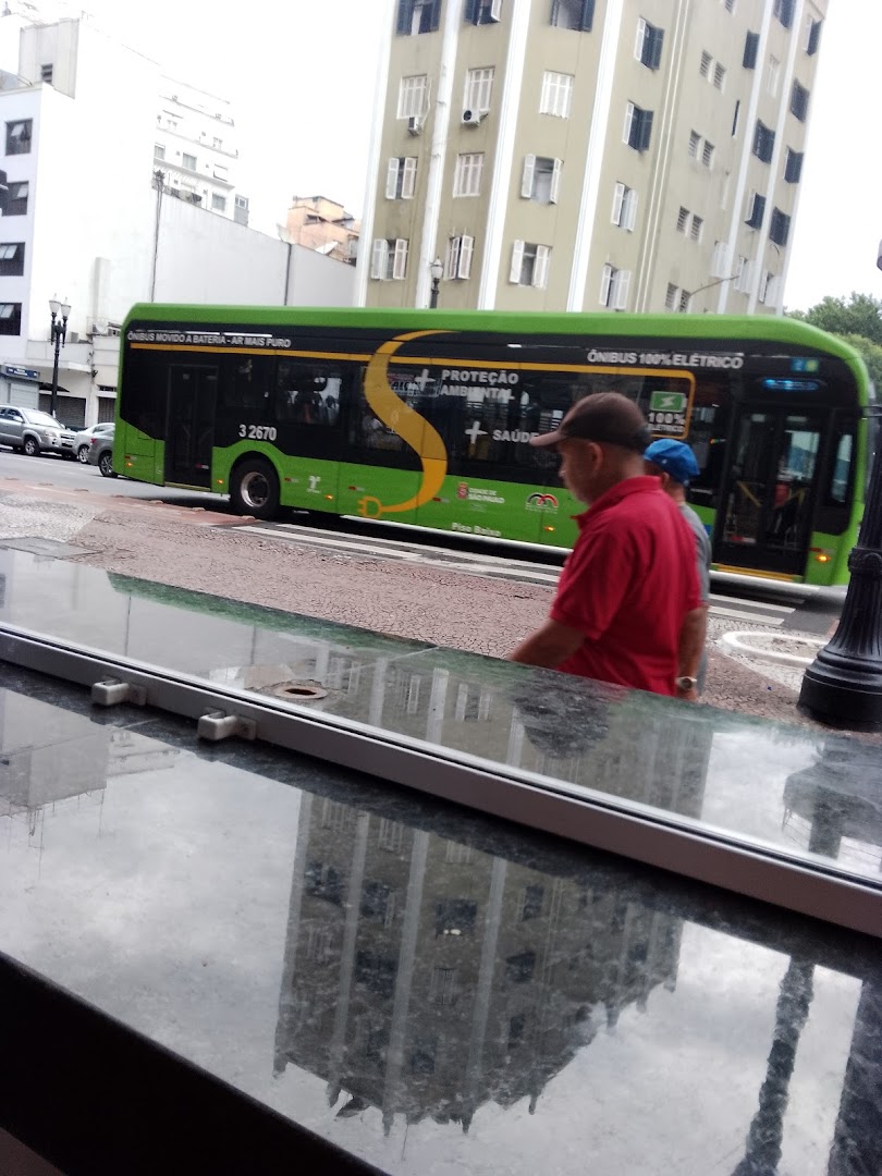A green electric bus with 'S' logo passes by people on a street with building reflections.