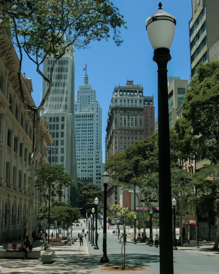 A street view with tall buildings, trees, lampposts, and people walking.
