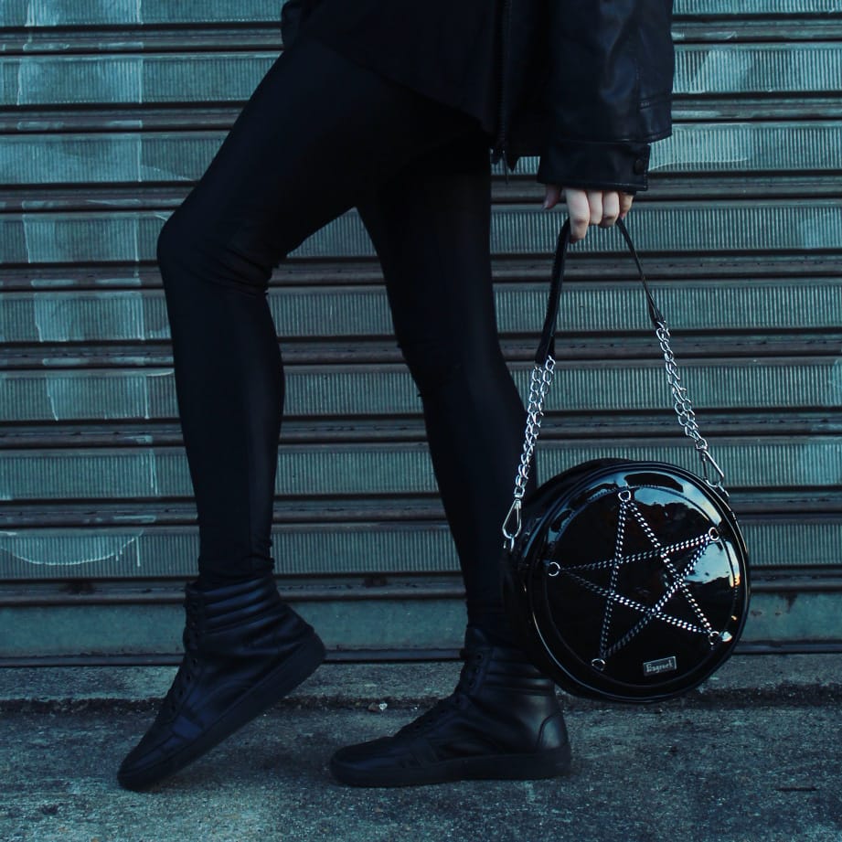 A person in black clothing and shoes holds a round black bag with a pentagram design.