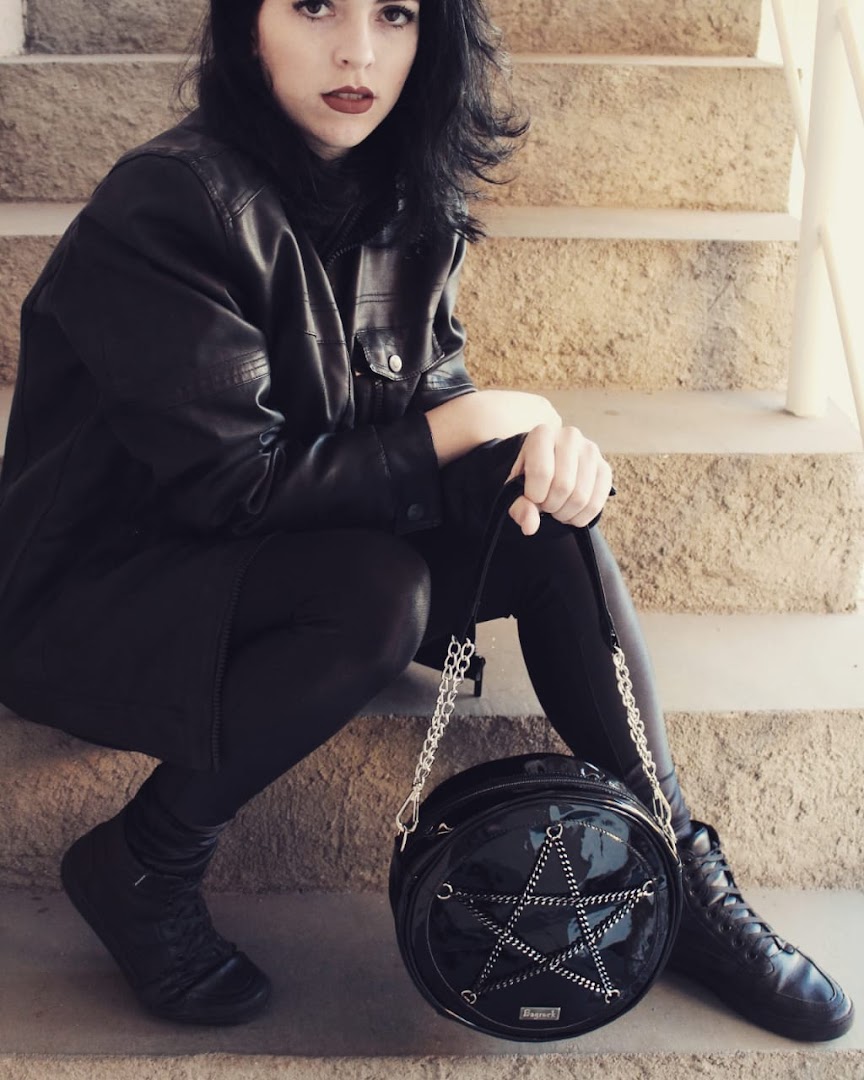 A person in black leather attire sits on stone steps, holding a circular black bag with a star design.