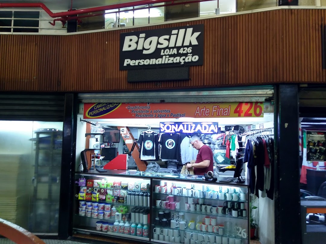 A shop called Bigsilk Personaliza displays custom t-shirts and mugs, with a staff member working behind the counter.