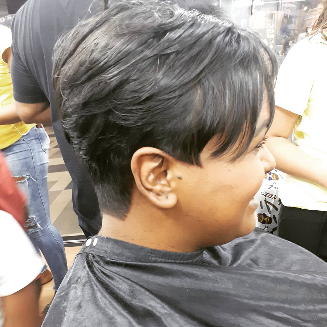 A person with a new haircut is smiling, wearing a black cape in a salon.
