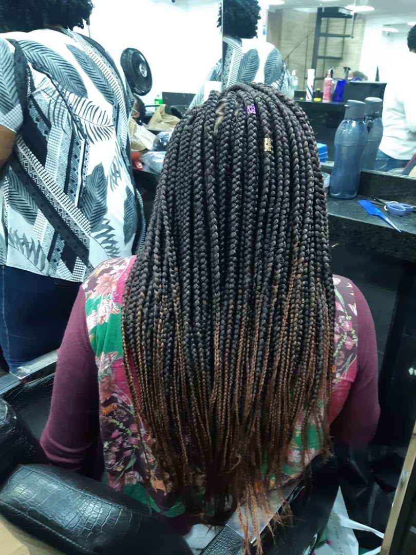 A person with long, braided hair sits in a salon chair, viewed from behind.