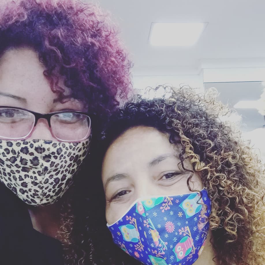 Two women with curly hair and face masks smiling at the camera.