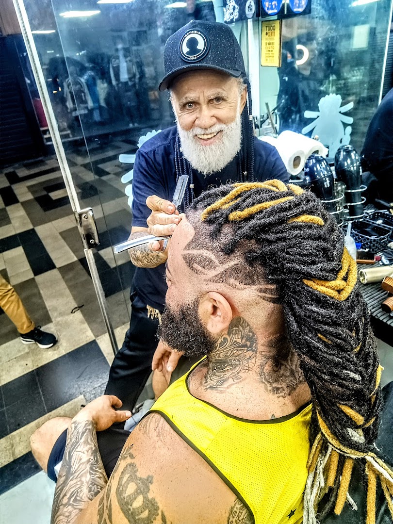 A barber with a white beard trims a client's hair with dreadlocks and intricate designs.