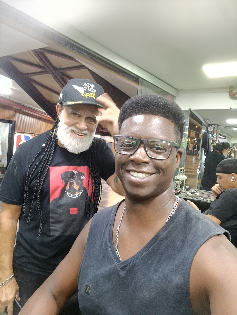 Two smiling men, one with a beard and braids, pose for a selfie inside a barbershop.