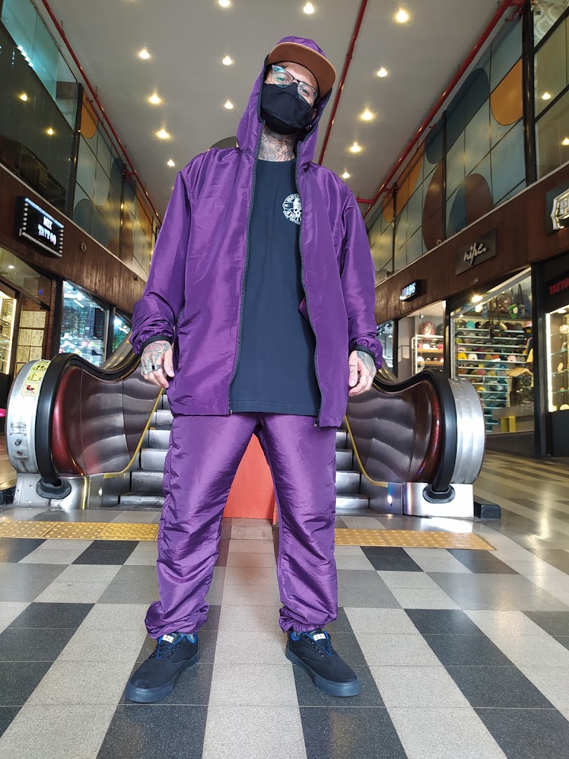A person wearing a purple tracksuit and mask stands in front of an escalator in a shopping mall.