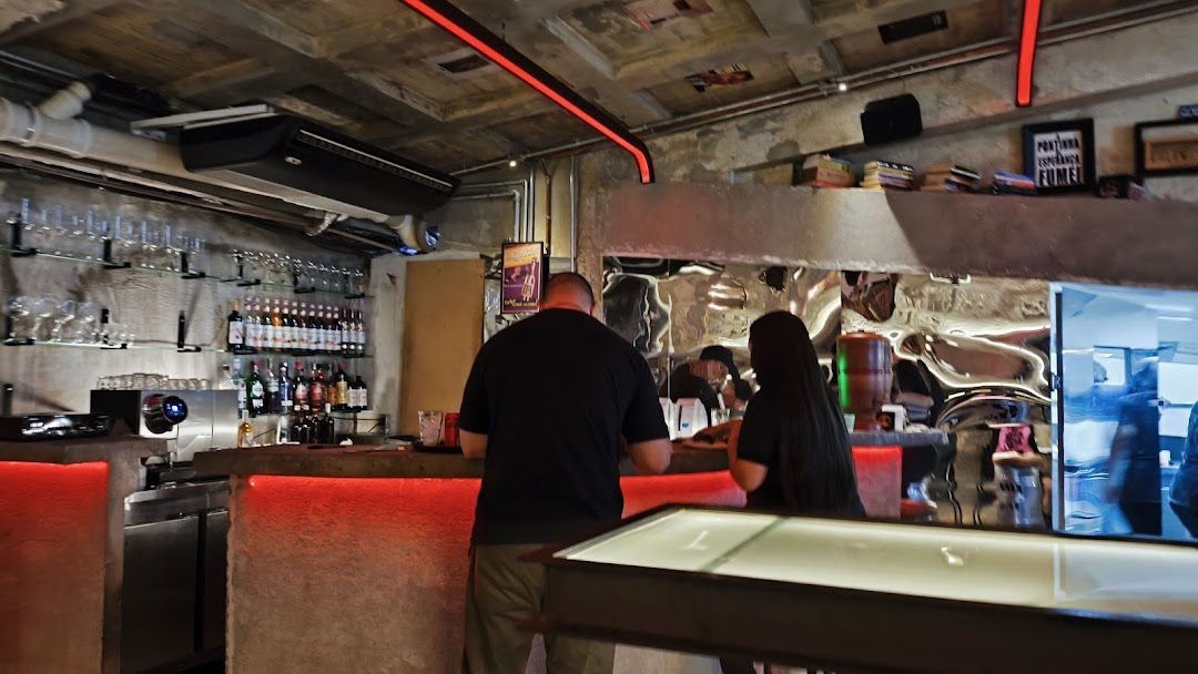 People at a bar with shelves of bottles and glasses, illuminated by red lights.