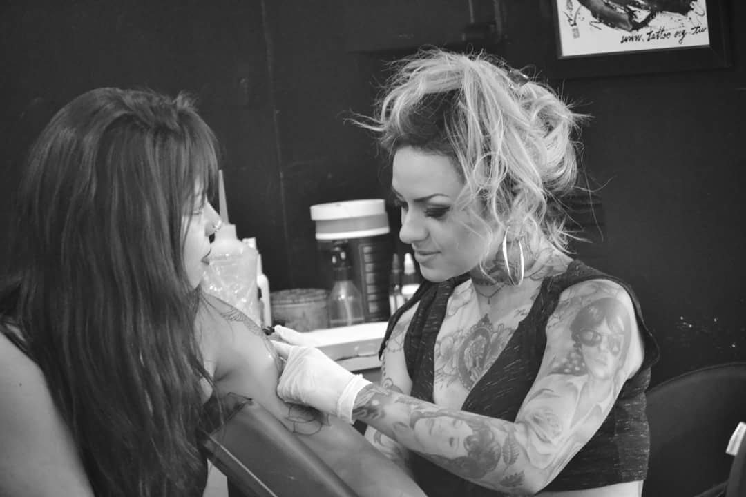 A tattoo artist works on a client's arm in a black and white studio.