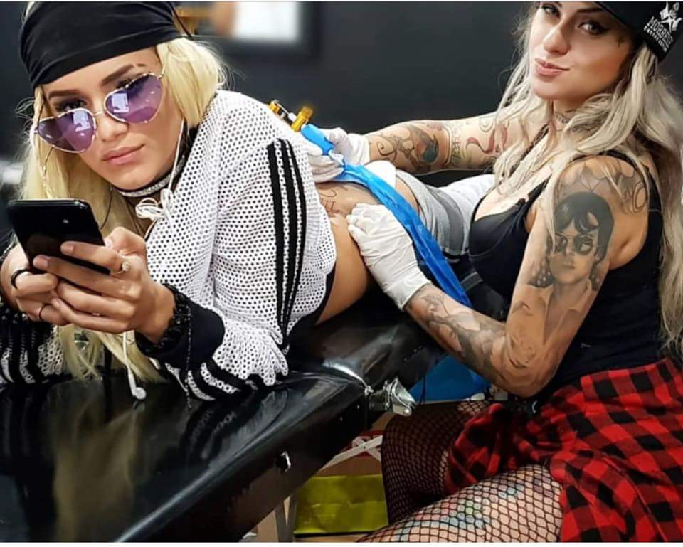 Two women in a tattoo studio, one getting tattooed while the other operates the tattoo machine.