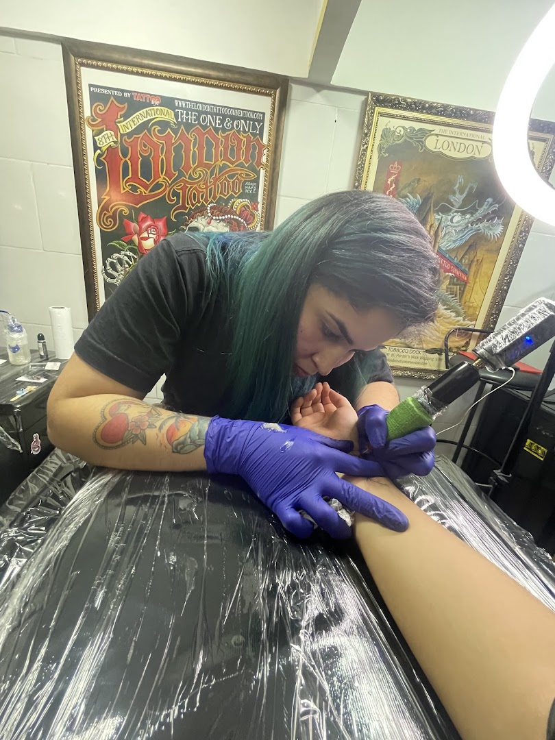 A tattoo artist with blue gloves inks a client's arm, with convention posters on the wall.