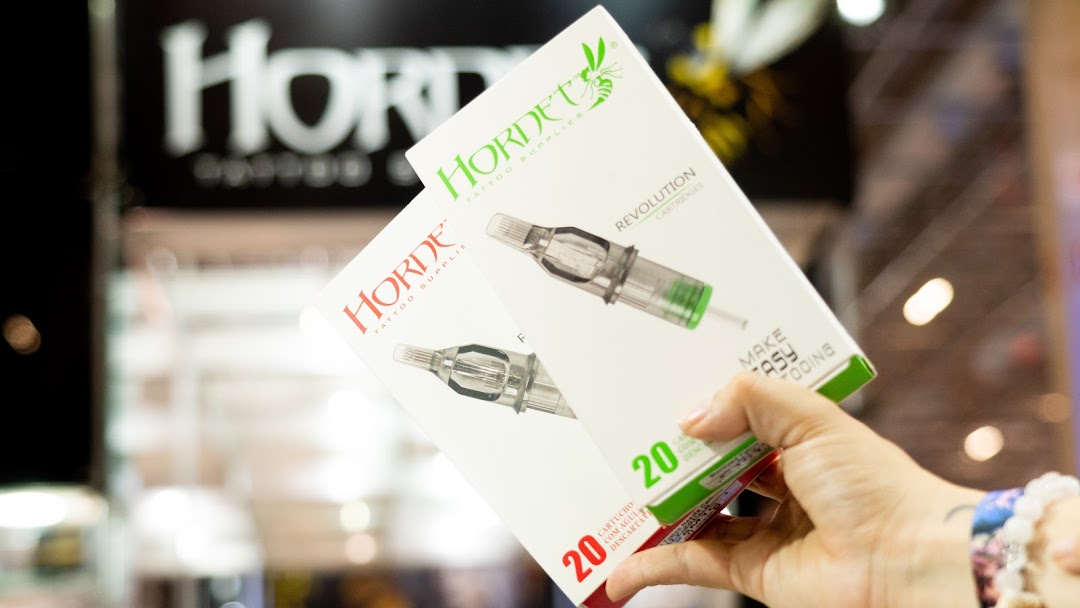 A hand holds two boxes of Hornet tattoo cartridges, with the shop's logo blurred in the background.