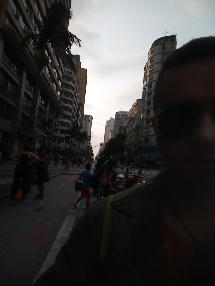 A person takes a selfie on a city street with buildings and people in the background.