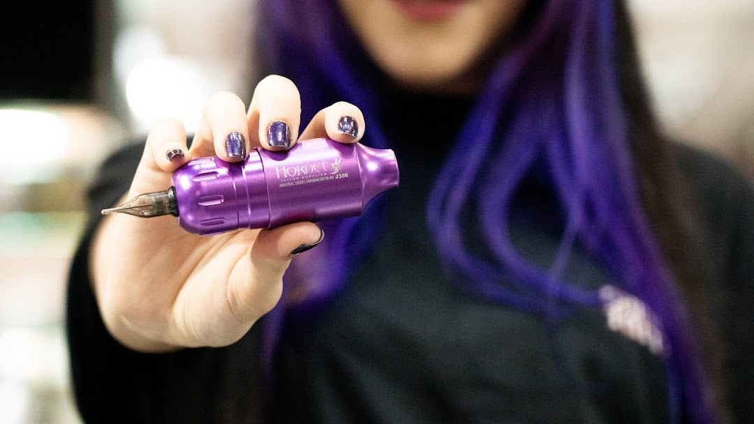 A person with purple hair holds a purple tattoo machine.