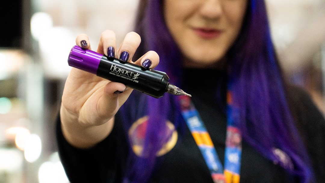 A person with purple hair holds a black and purple tattoo machine.