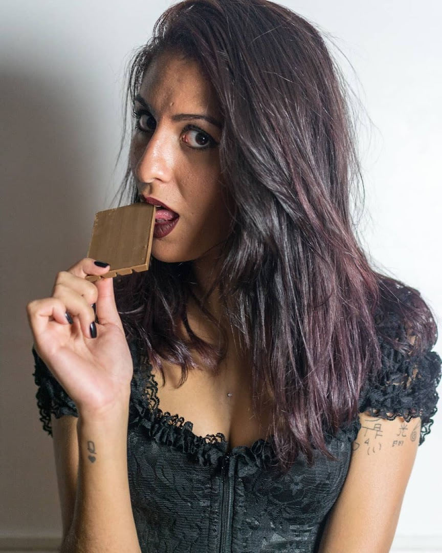 A woman with dark hair and dark lipstick bites into a square piece of chocolate.
