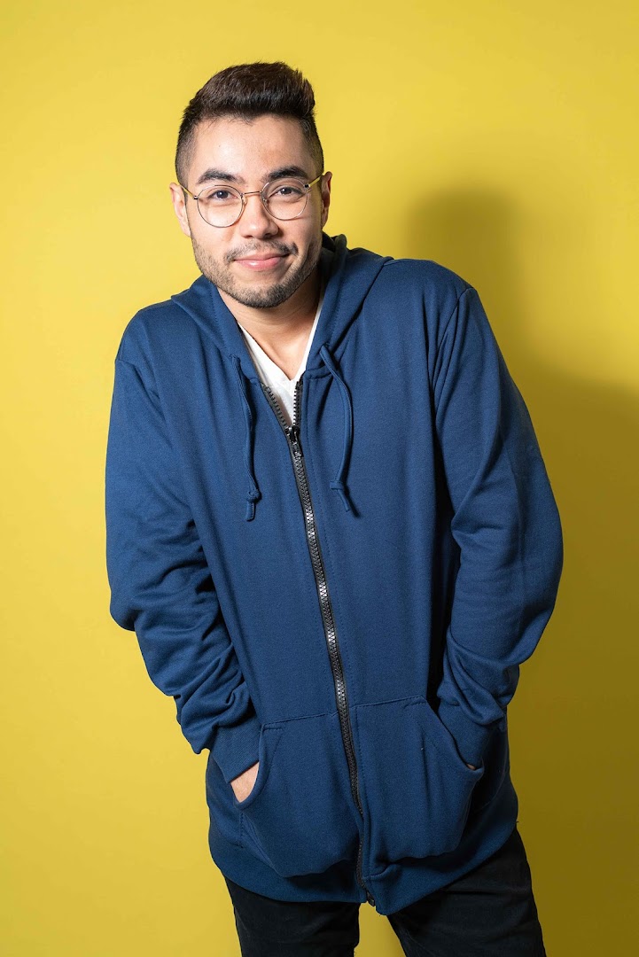 A man wearing a blue zip-up hoodie and glasses smiles against a yellow background.