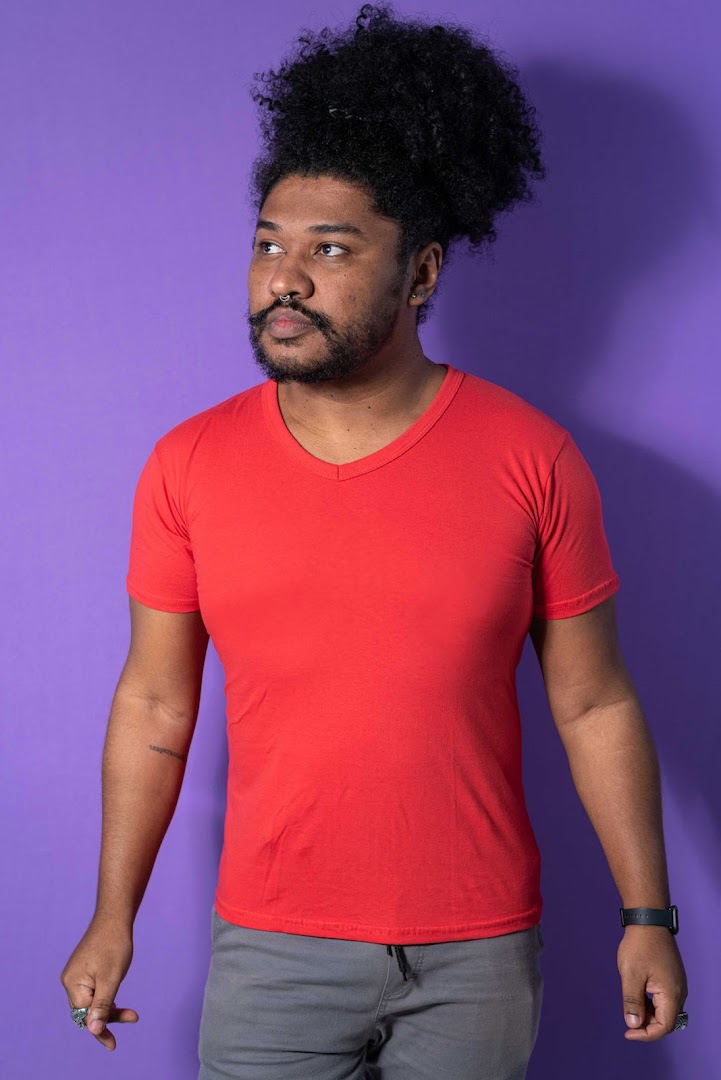 A man with curly hair and a beard wears a red V-neck t-shirt and grey pants.