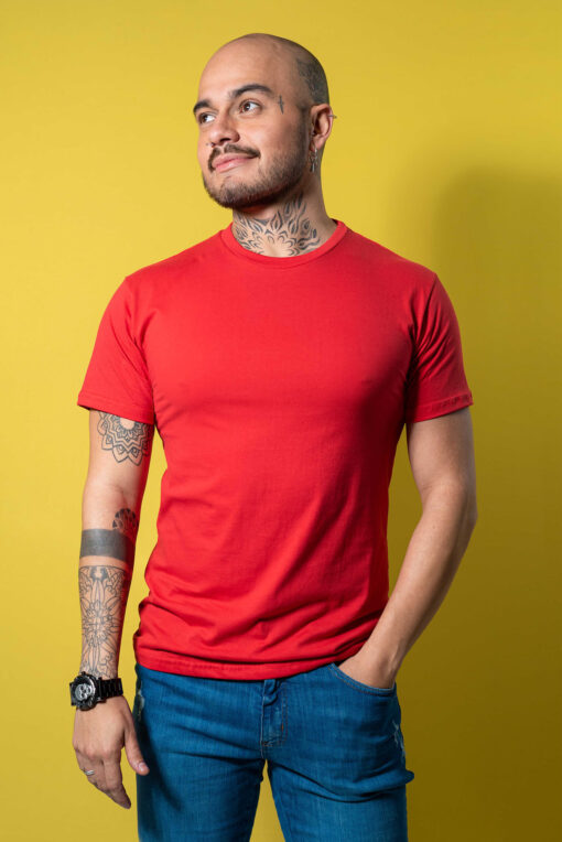 A man with tattoos wearing a red t-shirt and blue jeans against a yellow background.