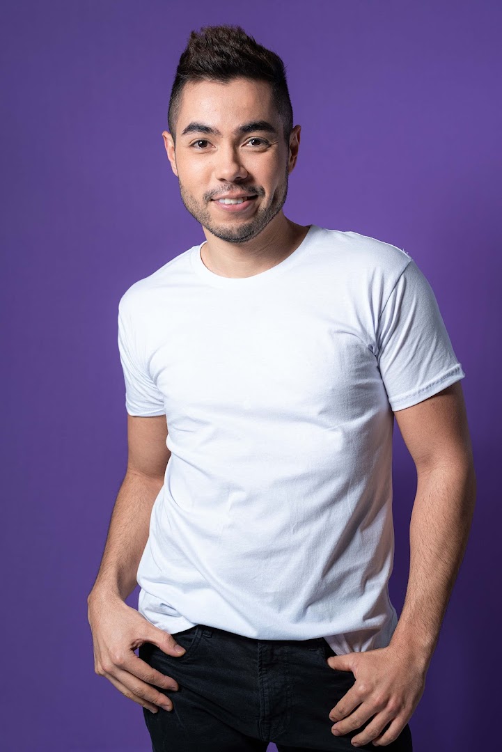 A smiling man with stylish hair and a white t-shirt poses against a purple background.