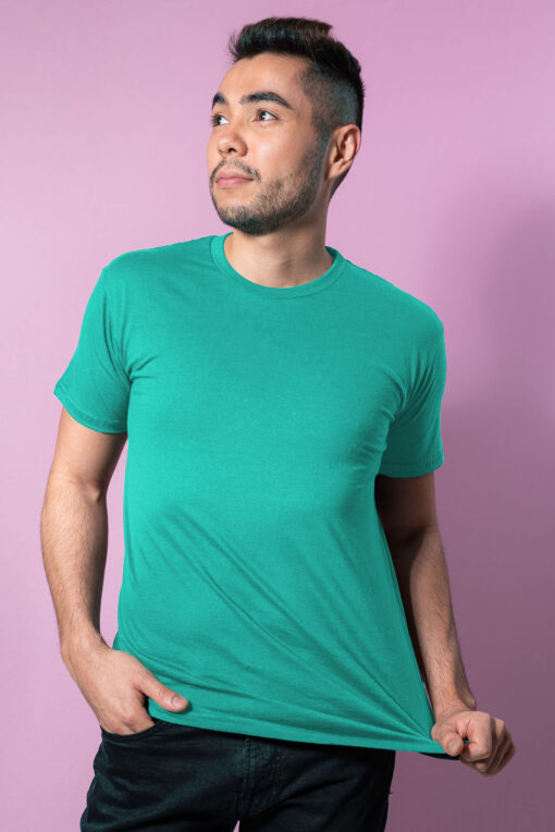 A man wearing a teal t-shirt and black jeans against a pink background.