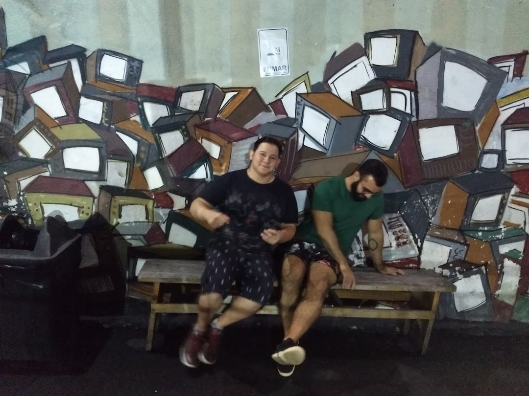Two men sit on a bench in front of a colorful mural of stacked televisions.