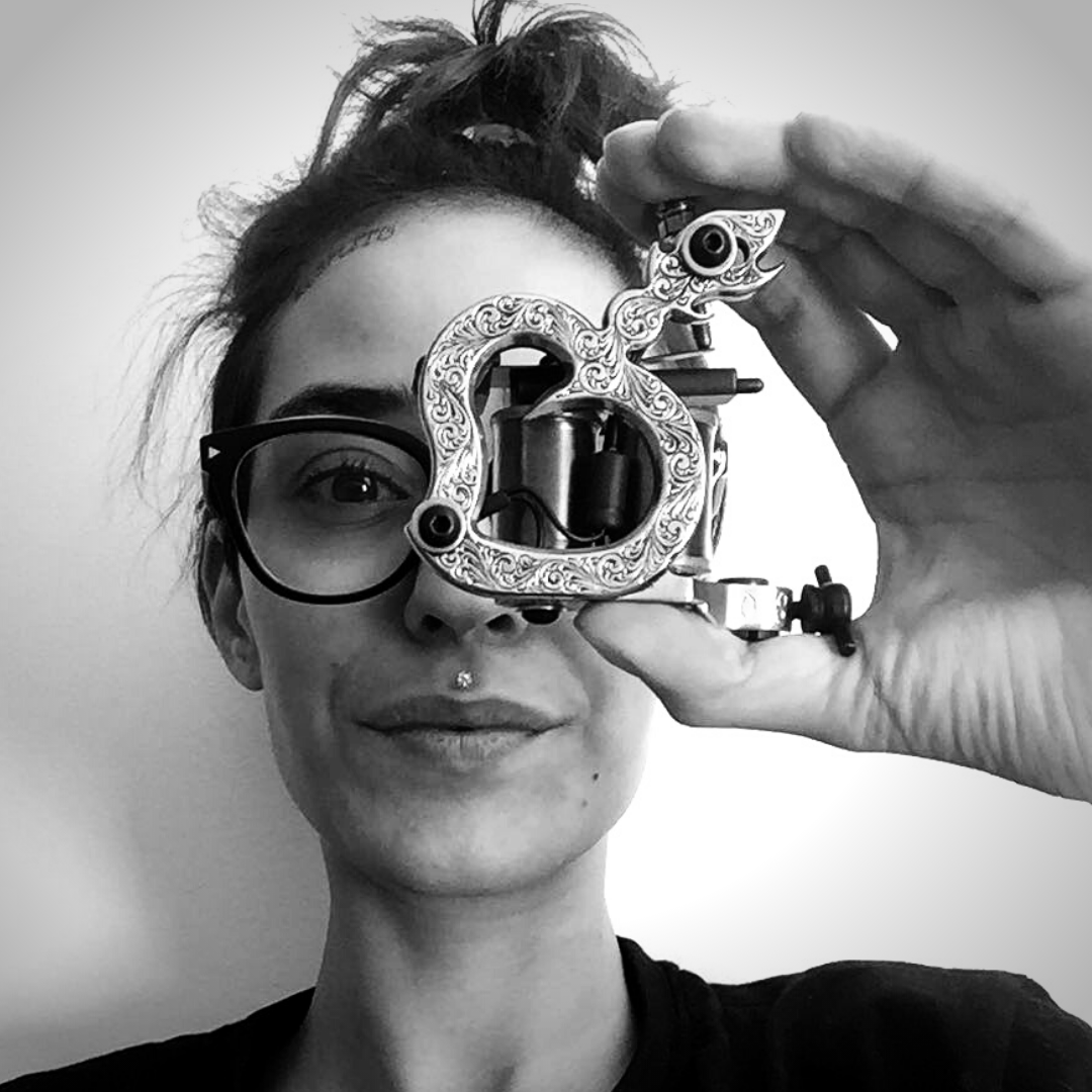 A person wearing glasses holds a tattoo machine up to their eye.