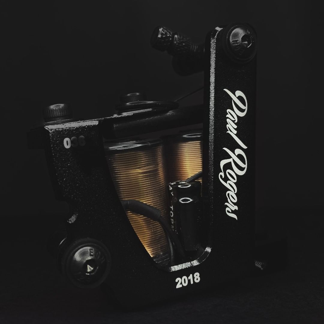 A black tattoo machine with gold coils, branded 'Paul Rogers', against a dark background.