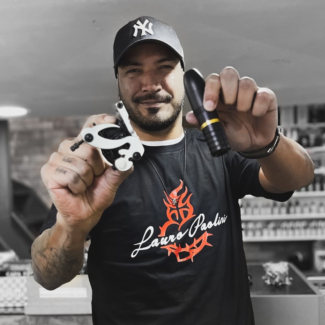 A man in a black t-shirt and baseball cap holds tattoo machines.