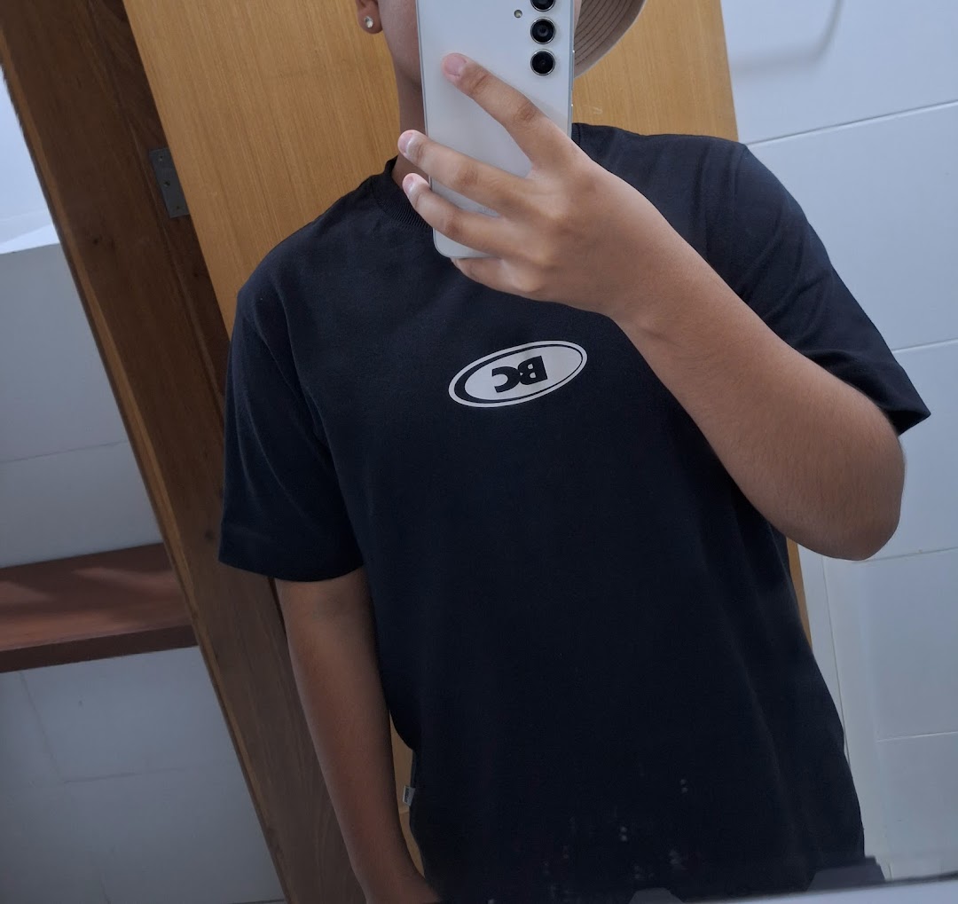 A person wearing a black t-shirt with a white oval logo takes a mirror selfie.