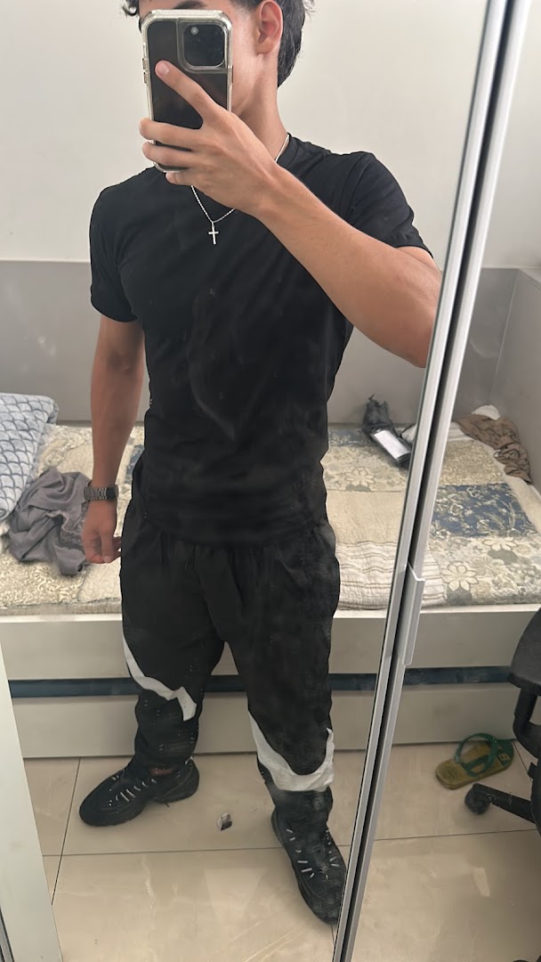 A person in a black t-shirt and black pants with white accents takes a mirror selfie.