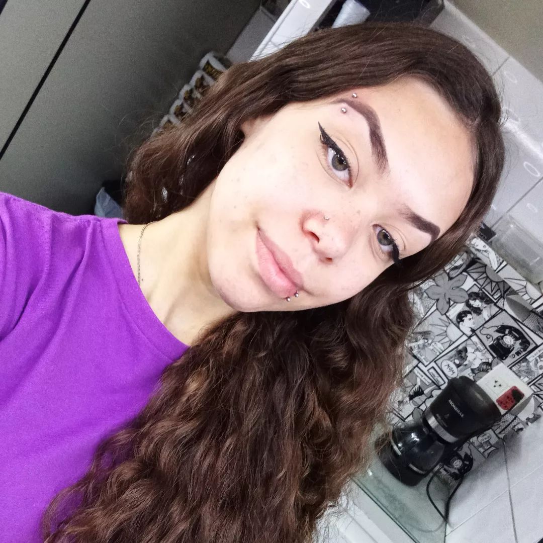 A young woman with long brown hair and multiple piercings smiles at the camera.