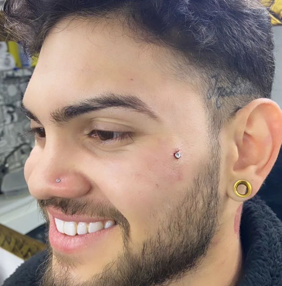 A man with a nose piercing, cheek piercing, and ear gauge smiles.