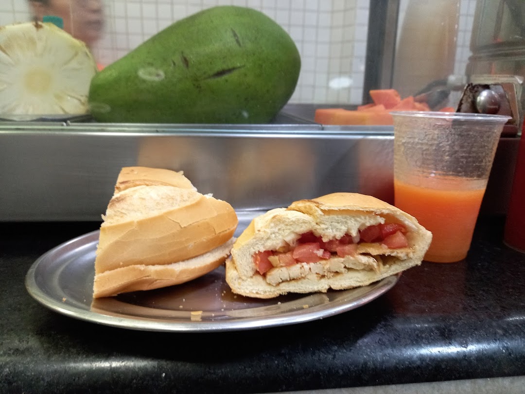 A sandwich and a glass of juice are displayed on a counter with fruits in the background.