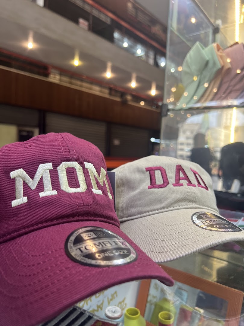 Close-up of two embroidered caps, one reading 'MOM' and the other 'DAD', displayed in a shop.