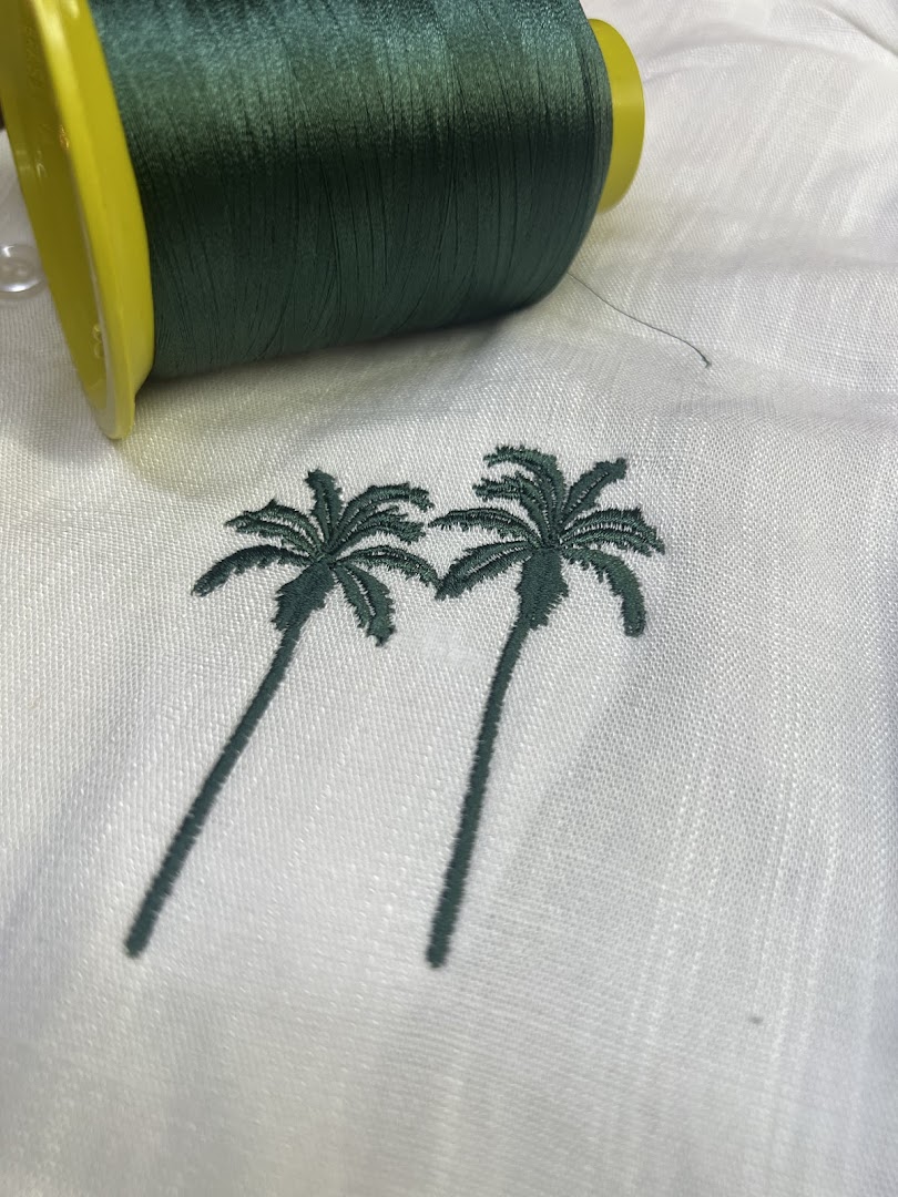 A spool of dark green thread rests on white fabric with embroidered palm trees.