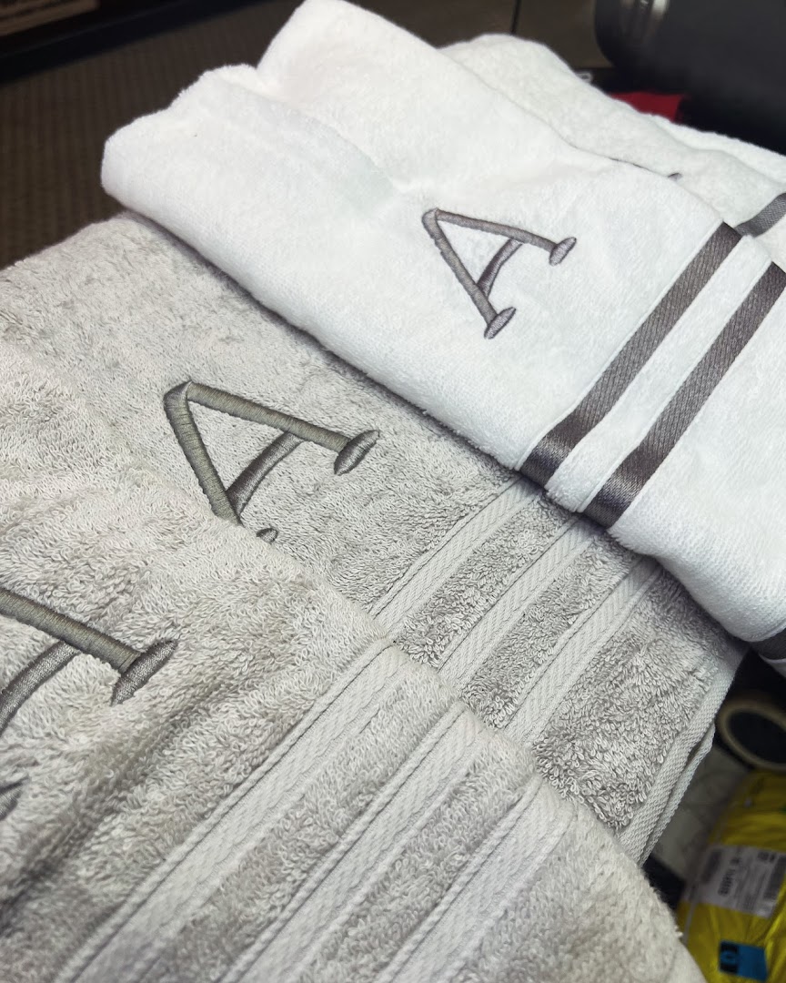 Close-up of two folded towels, one white and one grey, both embroidered with the letter 'A'.