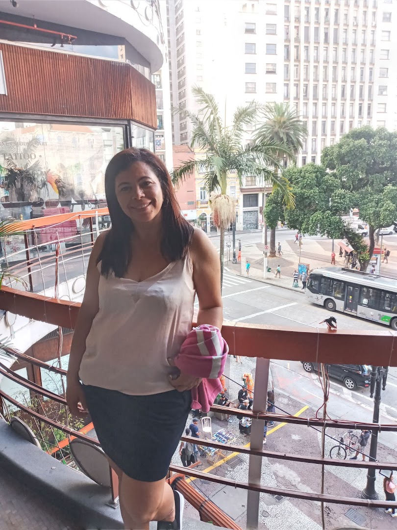 A woman stands on a balcony overlooking a busy city street with buildings and palm trees.