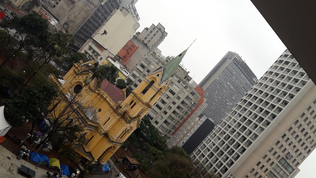 A yellow church with a green spire stands amidst tall city buildings.
