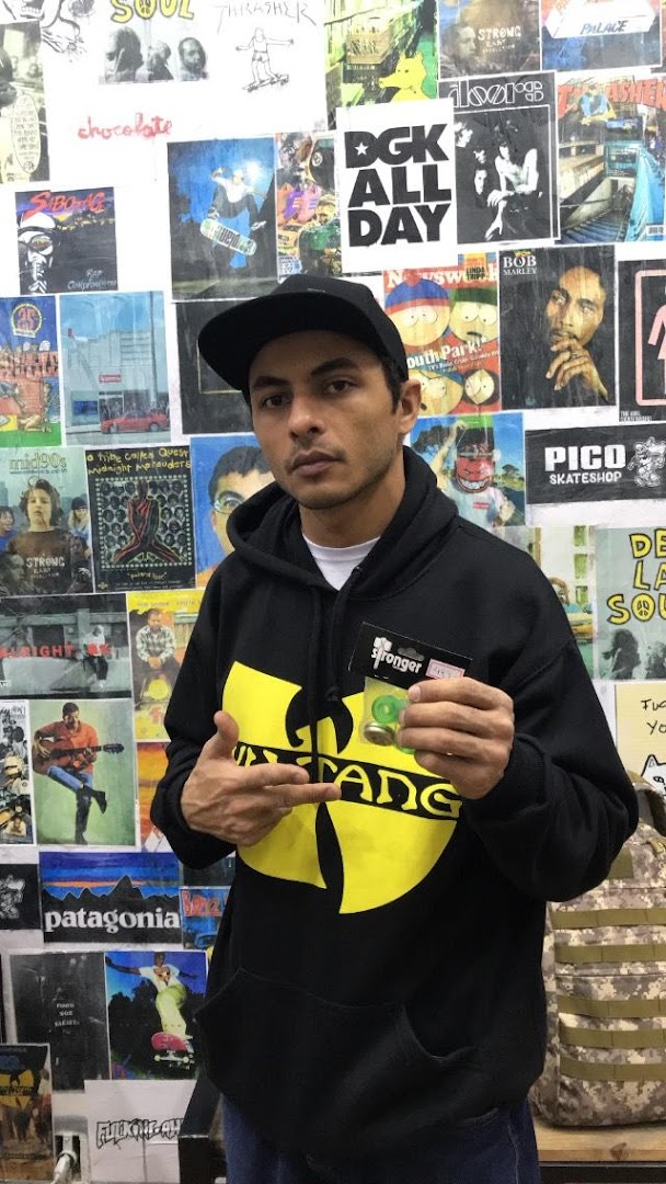 A man wearing a Wu-Tang Clan hoodie holds up a product in front of a wall covered in skate-related posters.