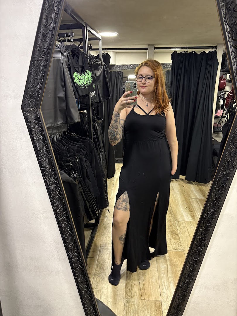 A woman in a black dress poses in front of a mirror, with racks of clothing visible.