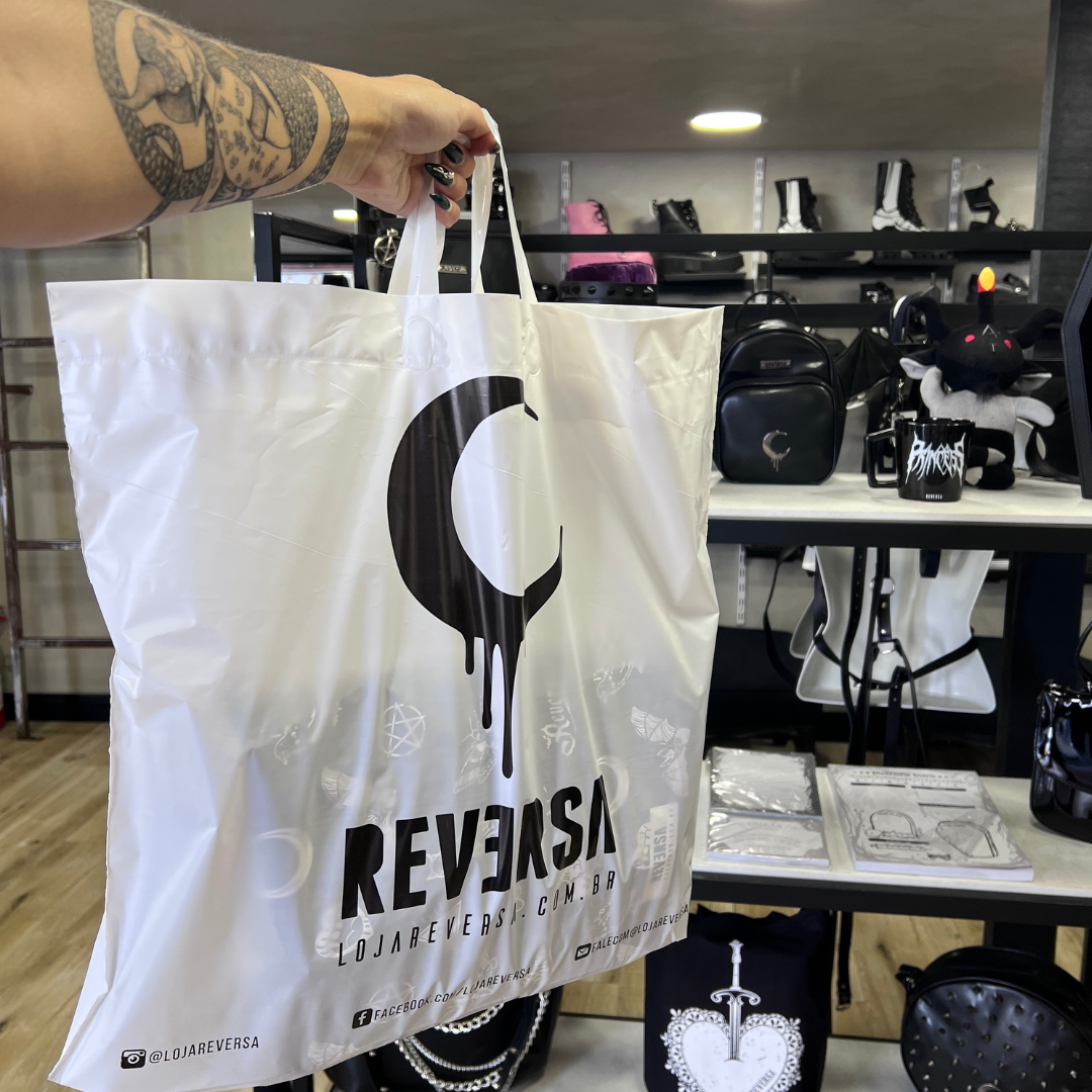 A hand holds a white shopping bag with the Reversa logo and various gothic-themed graphics.
