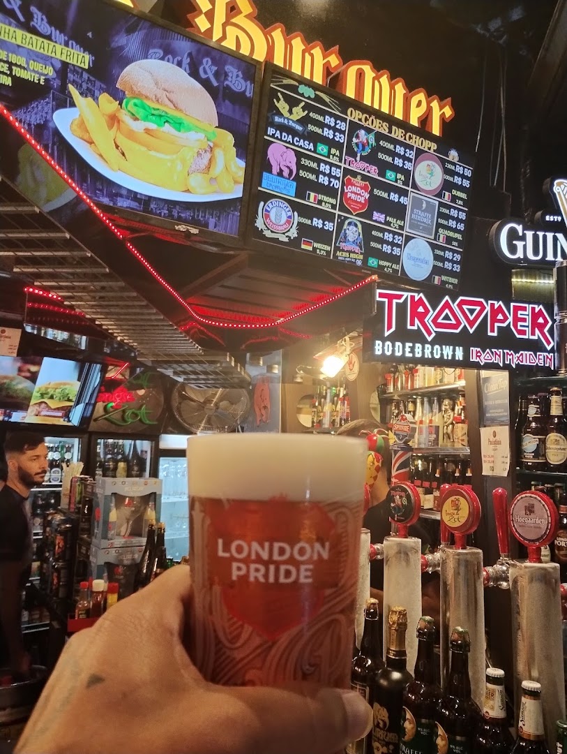A hand holds a glass of London Pride beer in front of a bar with a menu board and beer taps.