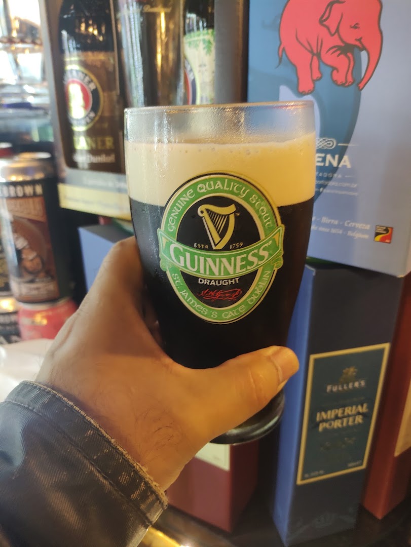 A hand holds a glass of Guinness beer with bottles and boxes in the background.