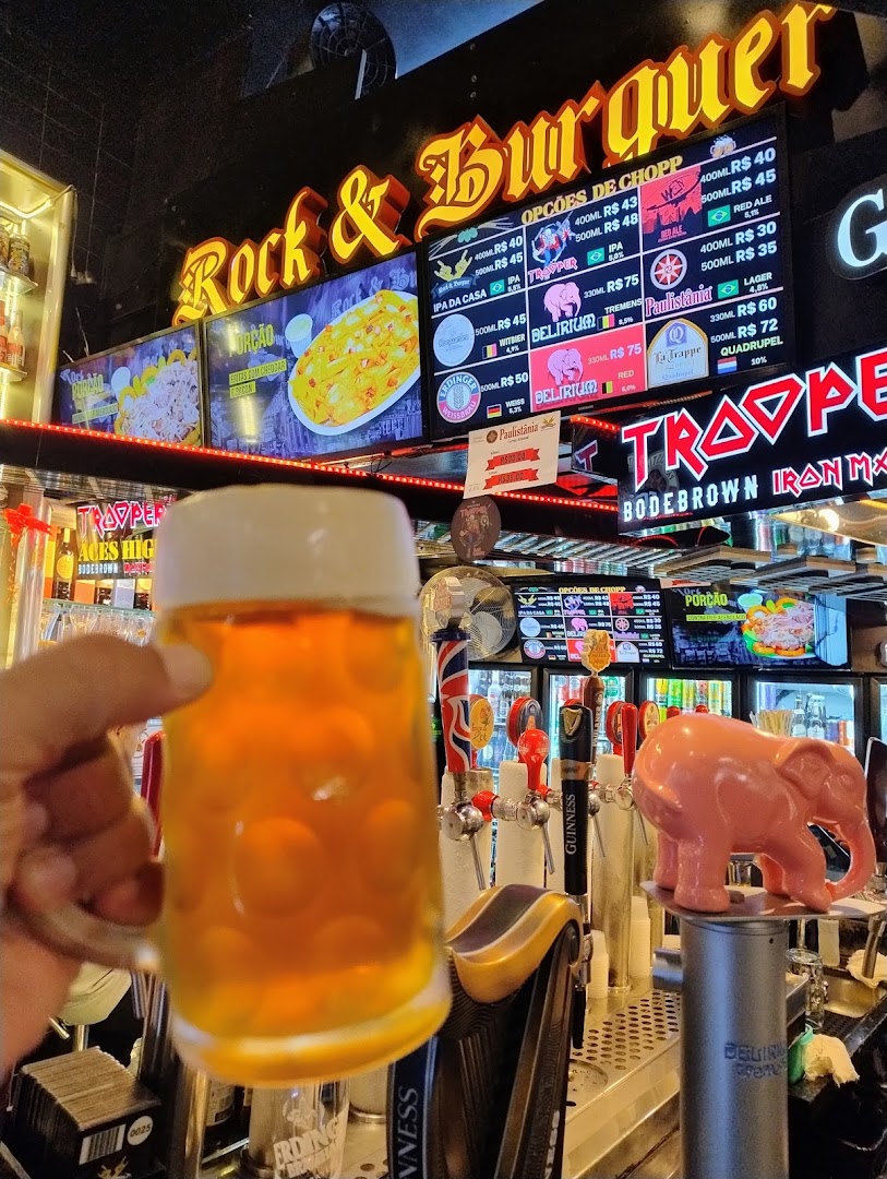 A hand holds a beer mug in front of a bar with beer taps and a menu board.