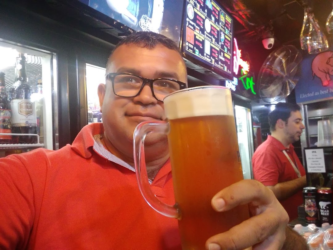 A man in a red shirt holds a large mug of beer in front of a bar.