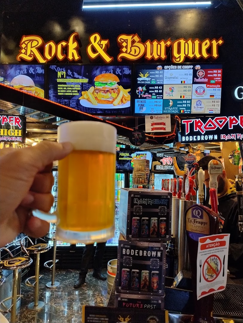A hand holds a beer mug in front of a Rock & Burguer restaurant with a menu and beer taps.